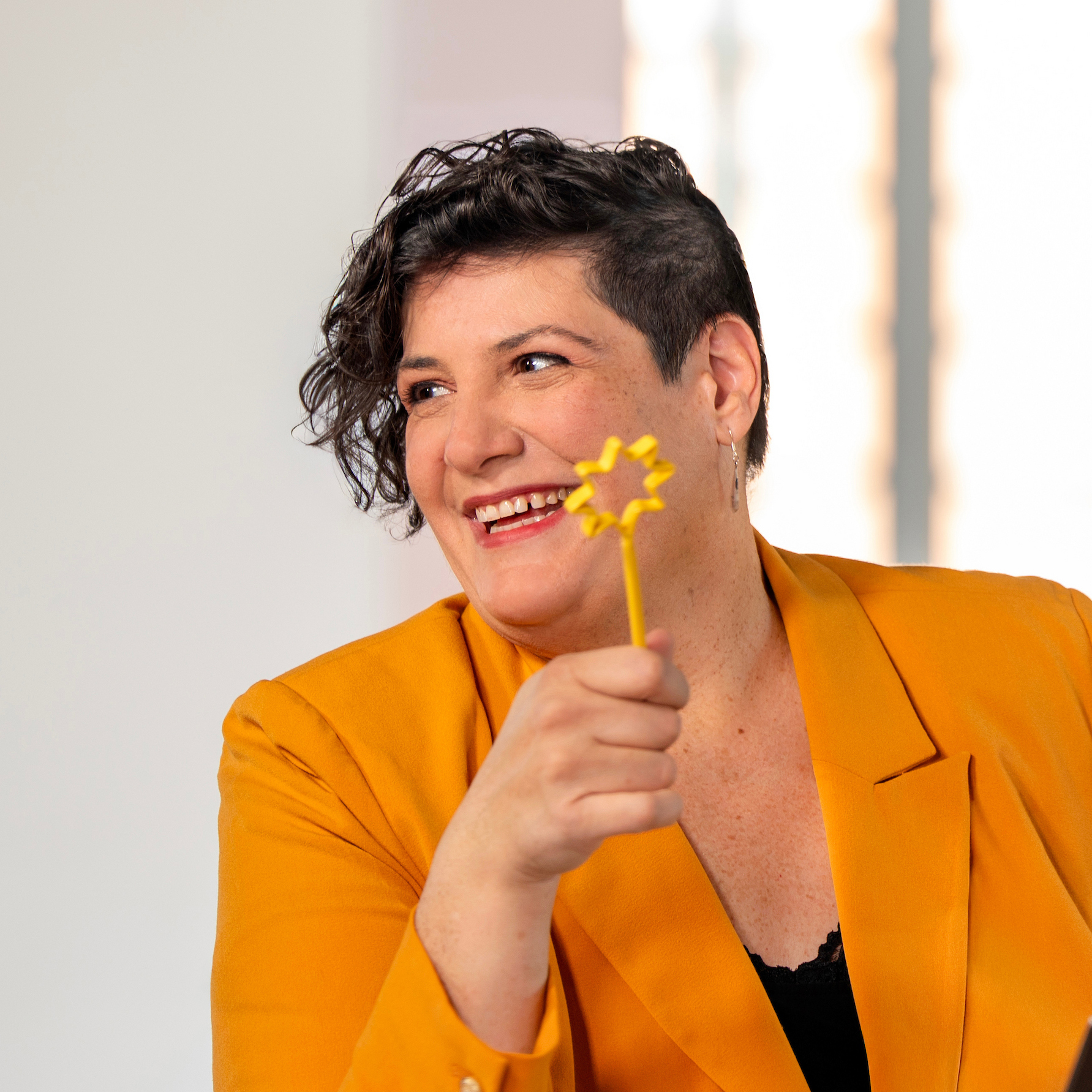 The Monarch Perri Collins in a gold blazer, sitting at a desk, holding a heart-shaped pen by her mouth